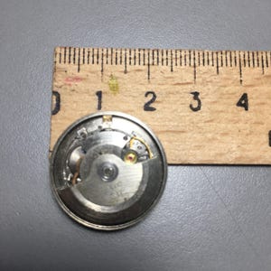 Puede incluir: Un movimiento de reloj desmontado con engranajes y componentes intrincados, colocado contra una regla de madera. El mecanismo del reloj tiene aproximadamente 2,5 cm de di&aacute;metro, mostrando sus detallados mecanismos internos. La regla proporciona una referencia de tama&ntilde;o.