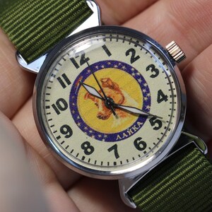 May include: A silver wristwatch with a white face and black hands. The face features a yellow and blue image of a dog with the word "LAIKA" printed below. The watch has a green nylon strap.