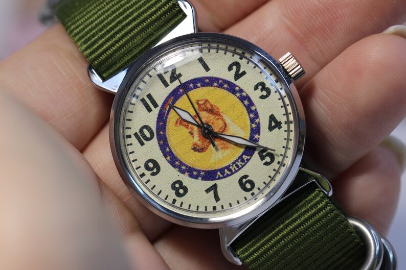 May include: A silver wristwatch with a white face and black hands. The face features a yellow and blue image of a dog with the word "LAIKA" printed below. The watch has a green nylon strap.
