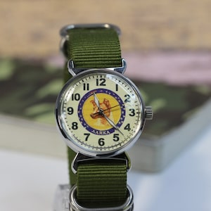 May include: A silver wristwatch with a white face and black numbers. The face of the watch features a yellow circle with a blue star and a picture of a dog with the text "LAIKA" underneath. The watch has a green nylon strap.