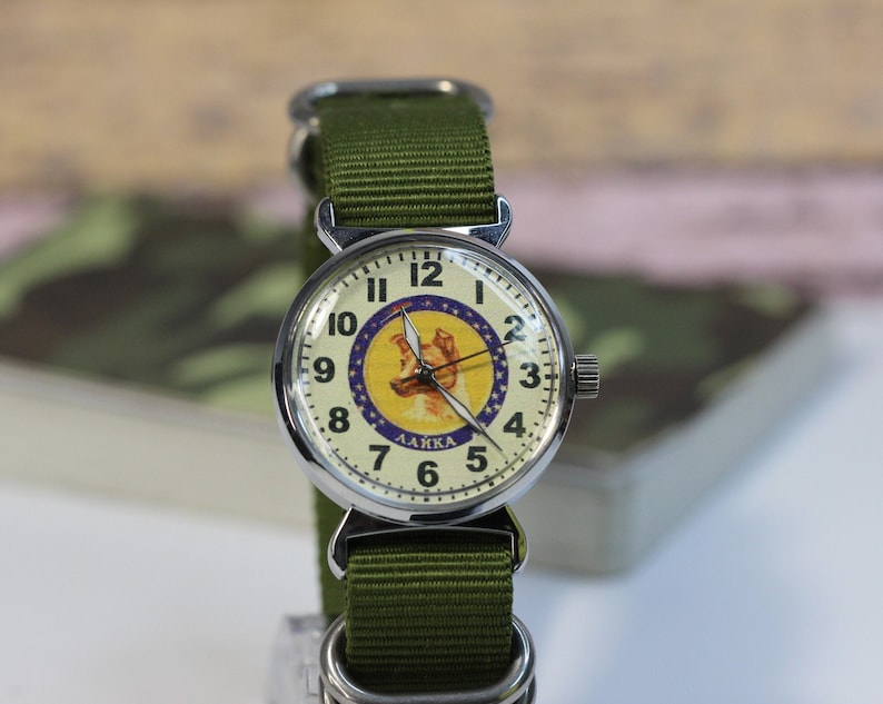 May include: A silver wristwatch with a white face and black numbers. The face of the watch features a yellow circle with a blue star and a picture of a dog with the text "LAIKA" underneath. The watch has a green nylon strap.