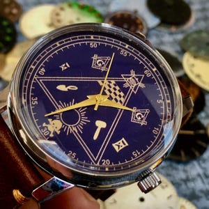 May include: A silver wristwatch with a dark blue face featuring gold-colored hands and symbols. The watch has a brown leather band. The background includes watch faces in various colors.
