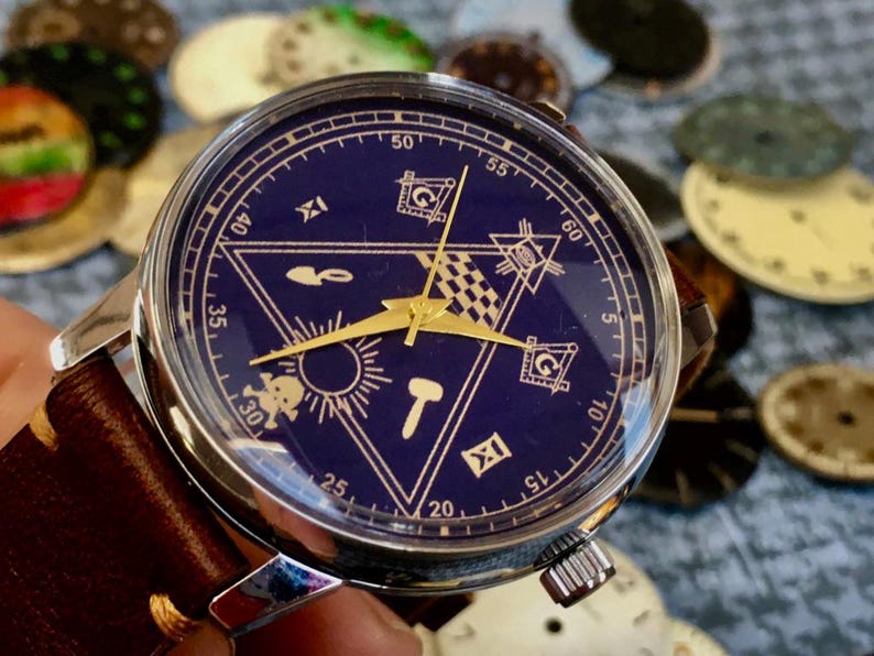 May include: A silver wristwatch with a dark blue face featuring gold-colored hands and symbols. The watch has a brown leather band. The background includes watch faces in various colors.
