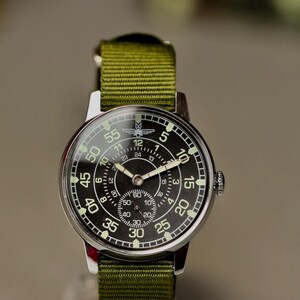 May include: A silver wristwatch with a black face and green canvas band. The watch face has white numbers and markings, and a small sub-dial at the center. The watch has a military-style design.