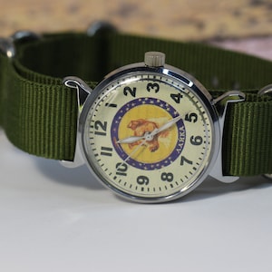 May include: A silver wristwatch with a white face and black numbers. The watch face features a yellow and purple image of a dog with the word "LAIKA" printed on it. The watch has a green nylon strap.