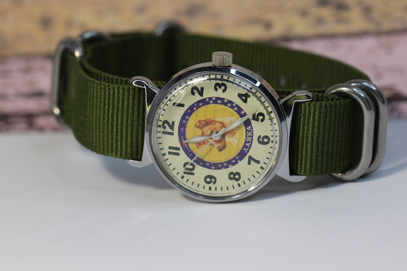 May include: A silver wristwatch with a white face and black numbers. The watch face features a yellow and purple image of a dog with the word "LAIKA" printed on it. The watch has a green nylon strap.