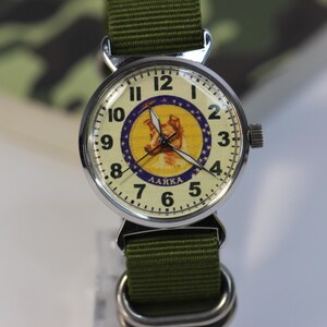 May include: A silver wristwatch with a green canvas band. The watch face has a white background with black numbers and hands. The center of the face features a yellow circle with a picture of a dog and the word "LAIKA" in Cyrillic letters.