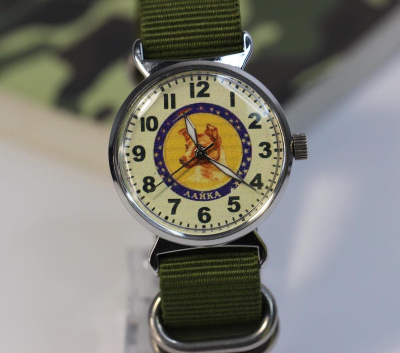 May include: A silver wristwatch with a green canvas band. The watch face has a white background with black numbers and hands. The center of the face features a yellow circle with a picture of a dog and the word "LAIKA" in Cyrillic letters.