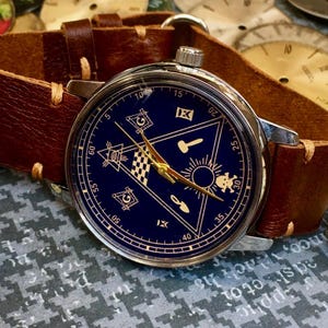 May include: A wristwatch with a dark blue face featuring gold hands and symbols, set in a silver case. The watch has a brown leather band. The watch face displays a triangle with symbols and the numbers 5, 10, 15, 20, 25, 30, 35, 40, 50, and 60.
