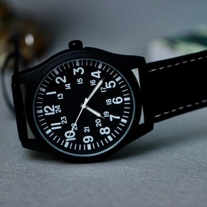 May include: Black wristwatch with a black face and white numerals. The watch has a black leather band with white stitching. The watch face displays the numbers 1 through 24. The watch is a classic, minimalist design.