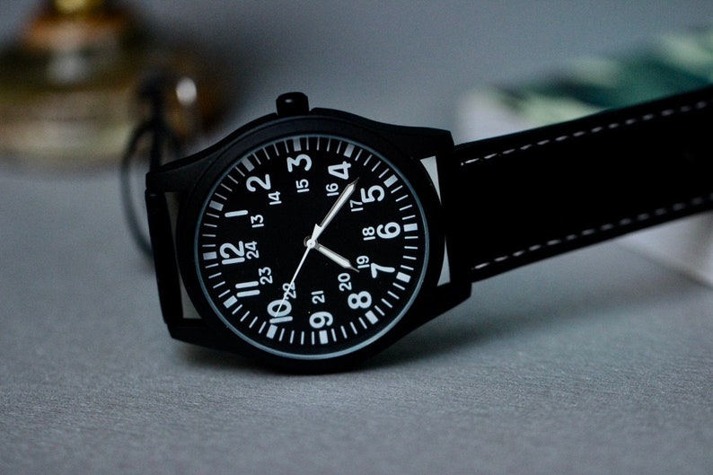 May include: Black wristwatch with a black face and white numerals. The watch has a black leather band with white stitching. The watch face displays the numbers 1 through 24. The watch is a classic, minimalist design.
