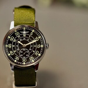 May include: A black and silver wristwatch with a green nylon strap. The watch face has a black background with white numbers and a small second hand dial. The watch has a white airplane logo at the top of the face.