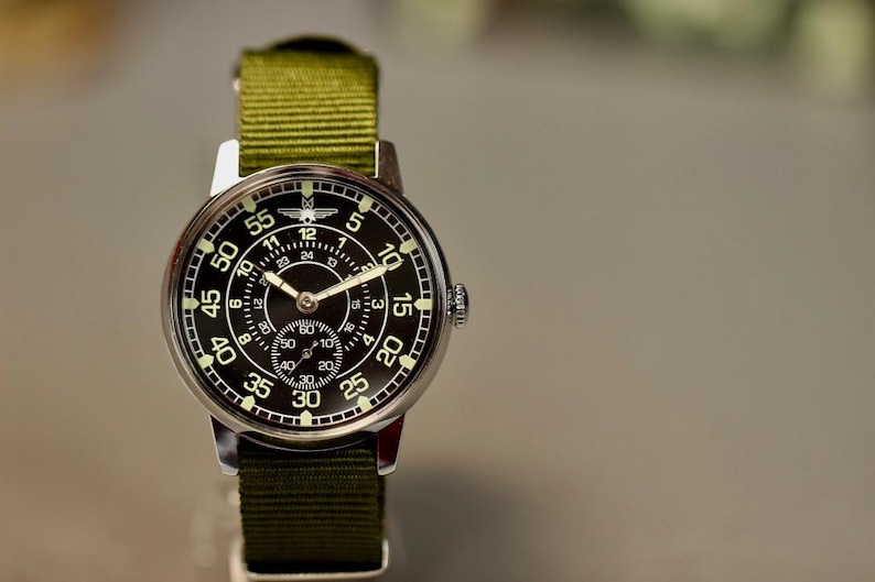 May include: A black and silver wristwatch with a green nylon strap. The watch face has a black background with white numbers and a small second hand dial. The watch has a white airplane logo at the top of the face.