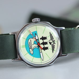 May include: A silver wristwatch with a green leather band. The watch face has a white background with a blue and green polar bear design and the text "БУРАН СССР".