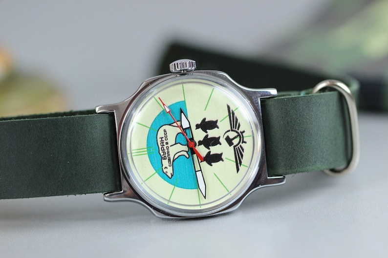 May include: A silver wristwatch with a green leather band. The watch face has a white background with a blue and green polar bear design and the text "БУРАН СССР".