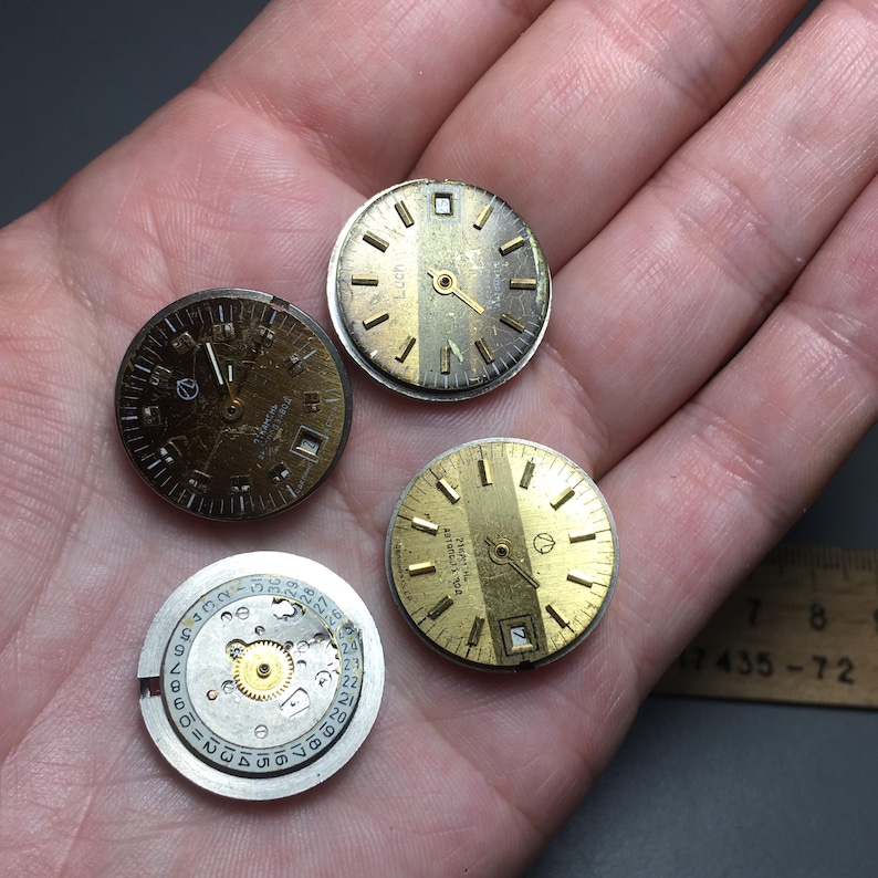 Puede incluir: Se muestran cuatro esferas de reloj vintage y un movimiento de reloj. Las esferas del reloj son doradas con marcas y manecillas negras. Una esfera de reloj tiene la palabra "Vostok" impresa. El movimiento del reloj es plateado con un engranaje dorado.