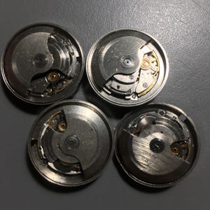 Puede incluir: Cuatro movimientos de reloj desmontados con engranajes, muelles y otros componentes internos visibles. Las piezas met&aacute;licas son plateadas, algunas con detalles dorados. Las piezas del reloj est&aacute;n dispuestas sobre una superficie gris.