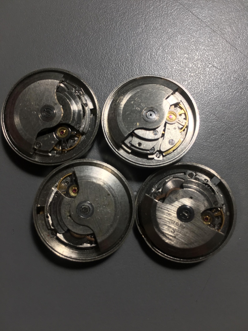 Puede incluir: Cuatro movimientos de reloj desmontados con engranajes, muelles y otros componentes internos visibles. Las piezas met&aacute;licas son plateadas, algunas con detalles dorados. Las piezas del reloj est&aacute;n dispuestas sobre una superficie gris.