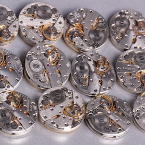 May include: A collection of 13 silver and gold watch movements with intricate gears and details.