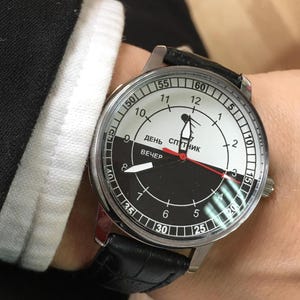 Puede incluir: Un reloj de pulsera con una correa de cuero negro y una caja plateada. La esfera del reloj está dividida en secciones negras y blancas, con el texto "ДЕНЬ СПУТНИК ВЕЧЕР" y números. Las manecillas son negras y rojas.