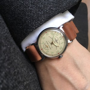 May include: A silver wristwatch with a brown leather band. The watch face is cream colored with black markings and features the text "ЮРИЙ ГАГАРИН" and "12 АПРЕЛЯ 1961" in Cyrillic script. The watch also has a small airplane logo.