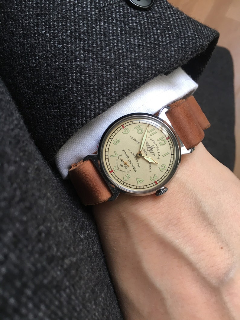 May include: A silver wristwatch with a brown leather band. The watch face is cream colored with black markings and features the text "ЮРИЙ ГАГАРИН" and "12 АПРЕЛЯ 1961" in Cyrillic script. The watch also has a small airplane logo.