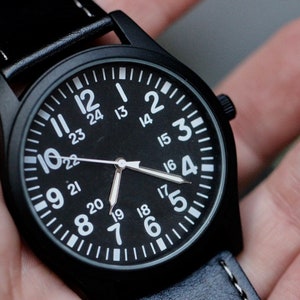 May include: A black wristwatch with a black face and white numerals. The watch has a black leather band with white stitching. The watch face displays the numbers 1 through 24. The hands are silver.