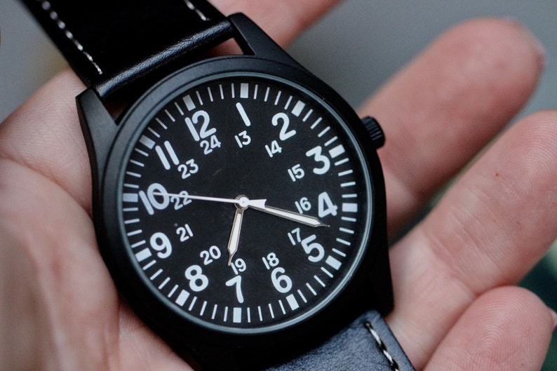 May include: A black wristwatch with a black face and white numerals. The watch has a black leather band with white stitching. The watch face displays the numbers 1 through 24. The hands are silver.