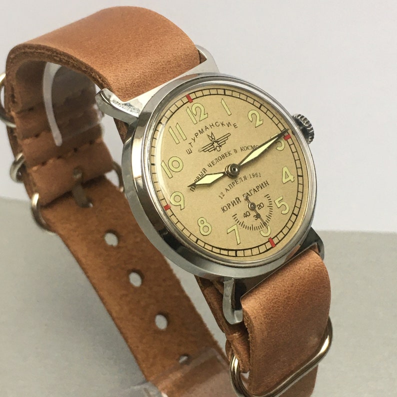 May include: A silver wristwatch with a tan leather band. The watch face is cream colored with black numbers and a red second hand. The watch face also has the text "ШТУРМАНСКИЕ" and "Первый Человек в Космос 12 Апреля 1961 ЮРИЙ ГАГАРИН".