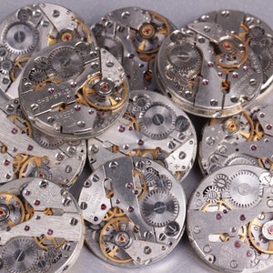 May include: A close-up of a pile of silver watch movements with intricate gears and mechanisms. The watch movements are arranged in a random pattern, with some overlapping.