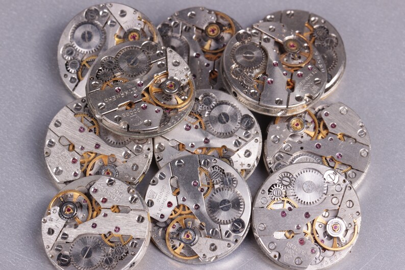 May include: A close-up of a pile of silver watch movements with intricate gears and mechanisms. The watch movements are arranged in a random pattern, with some overlapping.