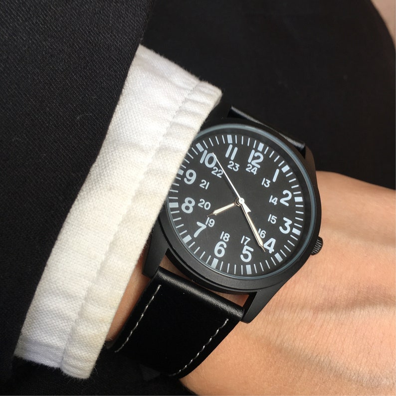 May include: A black wristwatch with a black face and white numerals. The watch has a black leather band with white stitching. The watch is being worn on a wrist, next to a white collared shirt.