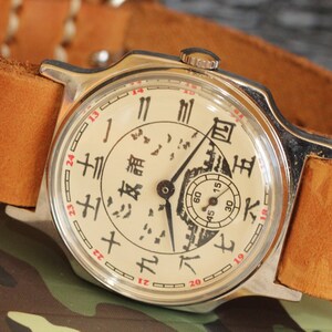 May include: A vintage wristwatch with a cream-colored face and black Chinese characters. The watch has a silver-colored case and a brown leather strap. The watch face features a small seconds dial and red numerals. The watch is set against a camouflage background.