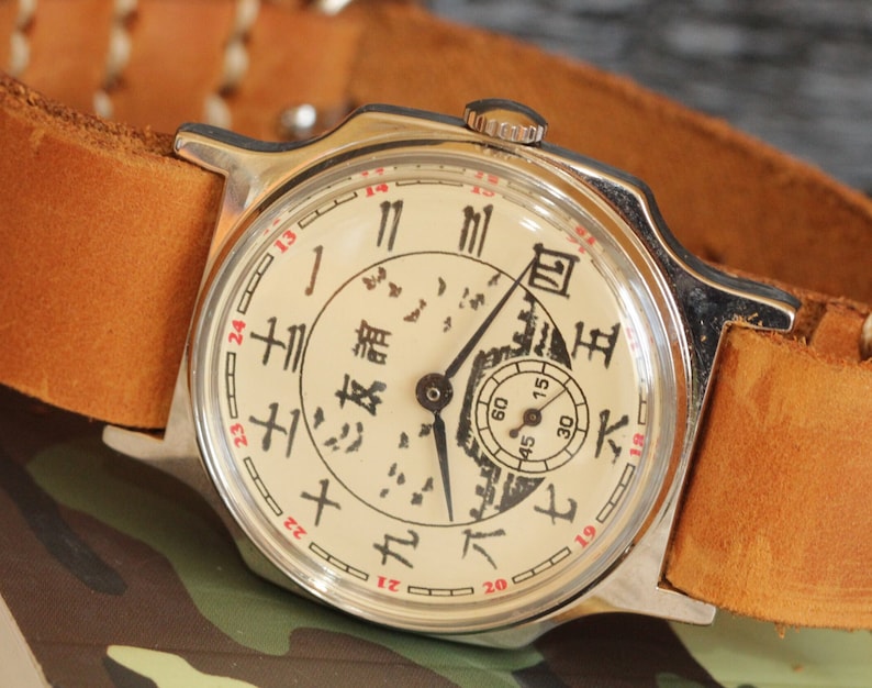 May include: A vintage wristwatch with a cream-colored face and black Chinese characters. The watch has a silver-colored case and a brown leather strap. The watch face features a small seconds dial and red numerals. The watch is set against a camouflage background.