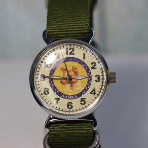May include: A silver wristwatch with a green nylon band. The watch face has a white background with a purple and yellow image of a dog, the text "ЛАЙКА" and black Roman numeral hour markers.