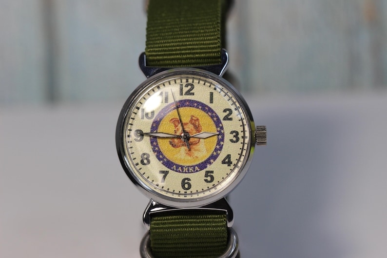 May include: A silver wristwatch with a green nylon band. The watch face has a white background with a purple and yellow image of a dog, the text "ЛАЙКА" and black Roman numeral hour markers.