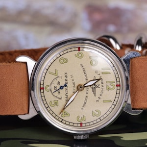 May include: A vintage wristwatch with a cream-colored face and black hands. The watch face has the text "ШТУРМАНСКИЕ" and "ПЕРВЫЙ ЧЕЛОВЕК В КОСМОСЕ" in Cyrillic script. The watch has a brown leather strap.