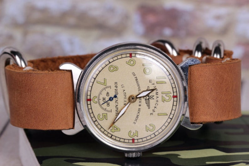 May include: A vintage wristwatch with a cream-colored face and black hands. The watch face has the text "ШТУРМАНСКИЕ" and "ПЕРВЫЙ ЧЕЛОВЕК В КОСМОСЕ" in Cyrillic script. The watch has a brown leather strap.