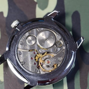 May include: Close-up of the back of a silver wristwatch with a mechanical movement. The watch face is visible through the clear case back, revealing the intricate gears and springs.