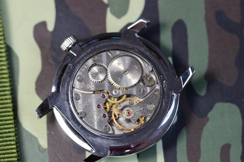 May include: Close-up of the back of a silver wristwatch with a mechanical movement. The watch face is visible through the clear case back, revealing the intricate gears and springs.