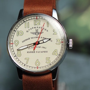 Puede incluir: Un reloj de pulsera plateado con una esfera blanca y números negros. La esfera del reloj tiene el texto "ШТУРМАНСКИЕ" y "ПЕРВЫЙ ЧЕЛОВЕК В КОСМОСЕ" con una aguja de segundos roja. El reloj tiene una correa de cuero marrón.