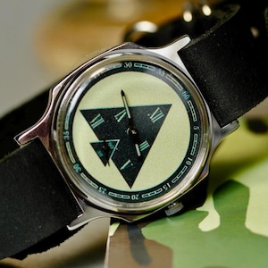 May include: A silver wristwatch with a black leather band. The watch face has a light green background with a black triangle design and Roman numeral hour markers. The watch is set against a camouflage pattern.