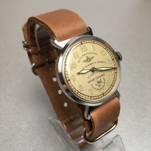 May include: A silver wristwatch with a brown leather band. The watch face is cream colored with black numbers and a red second hand. The watch face also has the text "ШТУРМАНСКИЕ" and "ПЕРВЫЙ ЧЕЛОВЕК В КОСМОСЕ" in Cyrillic script. The watch face also has the text "12 АПРЕЛЯ 1961" and "ЮРИЙ ГАГАРИН" in Cyrillic script.