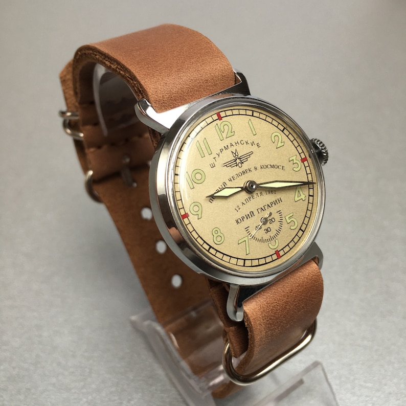 May include: A silver wristwatch with a brown leather band. The watch face is cream colored with black numbers and a red second hand. The watch face also has the text "ШТУРМАНСКИЕ" and "ПЕРВЫЙ ЧЕЛОВЕК В КОСМОСЕ" in Cyrillic script. The watch face also has the text "12 АПРЕЛЯ 1961" and "ЮРИЙ ГАГАРИН" in Cyrillic script.