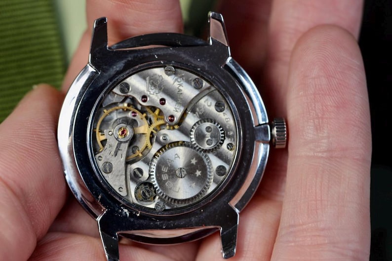 May include: Close-up of the back of a silver wristwatch with a mechanical movement. The watch face is visible through the clear case back, showing the intricate gears and springs.