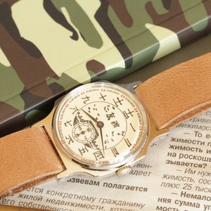 May include: A vintage wristwatch with a cream-colored face featuring black and red markings and a small subdial. The watch has a light brown leather strap and a gold-colored case. The background includes a camouflage pattern.