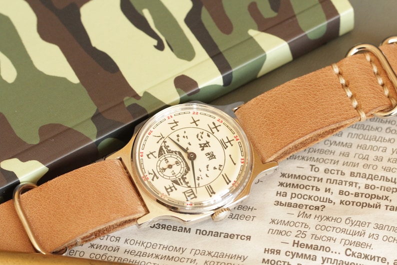 May include: A vintage wristwatch with a cream-colored face featuring black and red markings and a small subdial. The watch has a light brown leather strap and a gold-colored case. The background includes a camouflage pattern.