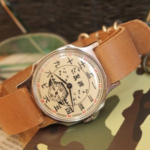 May include: A vintage wristwatch with a cream-colored face featuring black and red numerals and Chinese characters. The watch has a silver-colored case and a light brown leather strap. The watch face includes a small subdial and a detailed illustration.