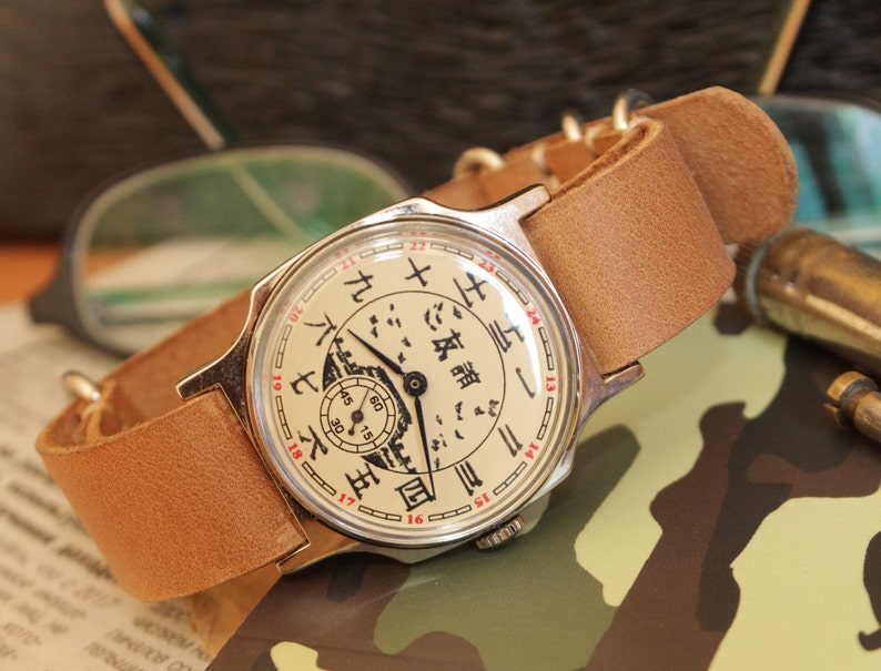 May include: A vintage wristwatch with a cream-colored face featuring black and red numerals and Chinese characters. The watch has a silver-colored case and a light brown leather strap. The watch face includes a small subdial and a detailed illustration.