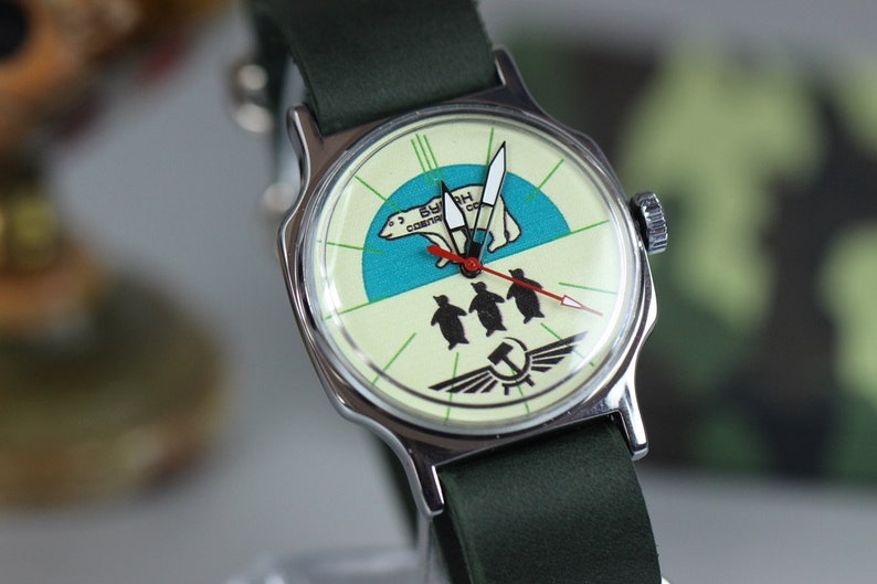 May include: A vintage wristwatch with a white face and a green strap. The watch face features a polar bear and penguins with the text "В.С.С.Р. СССР" in red.